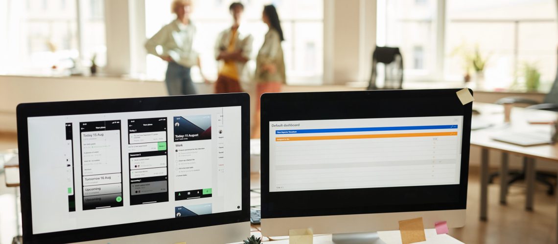 Image of computer monitors with new software on the table at office with people working in the background